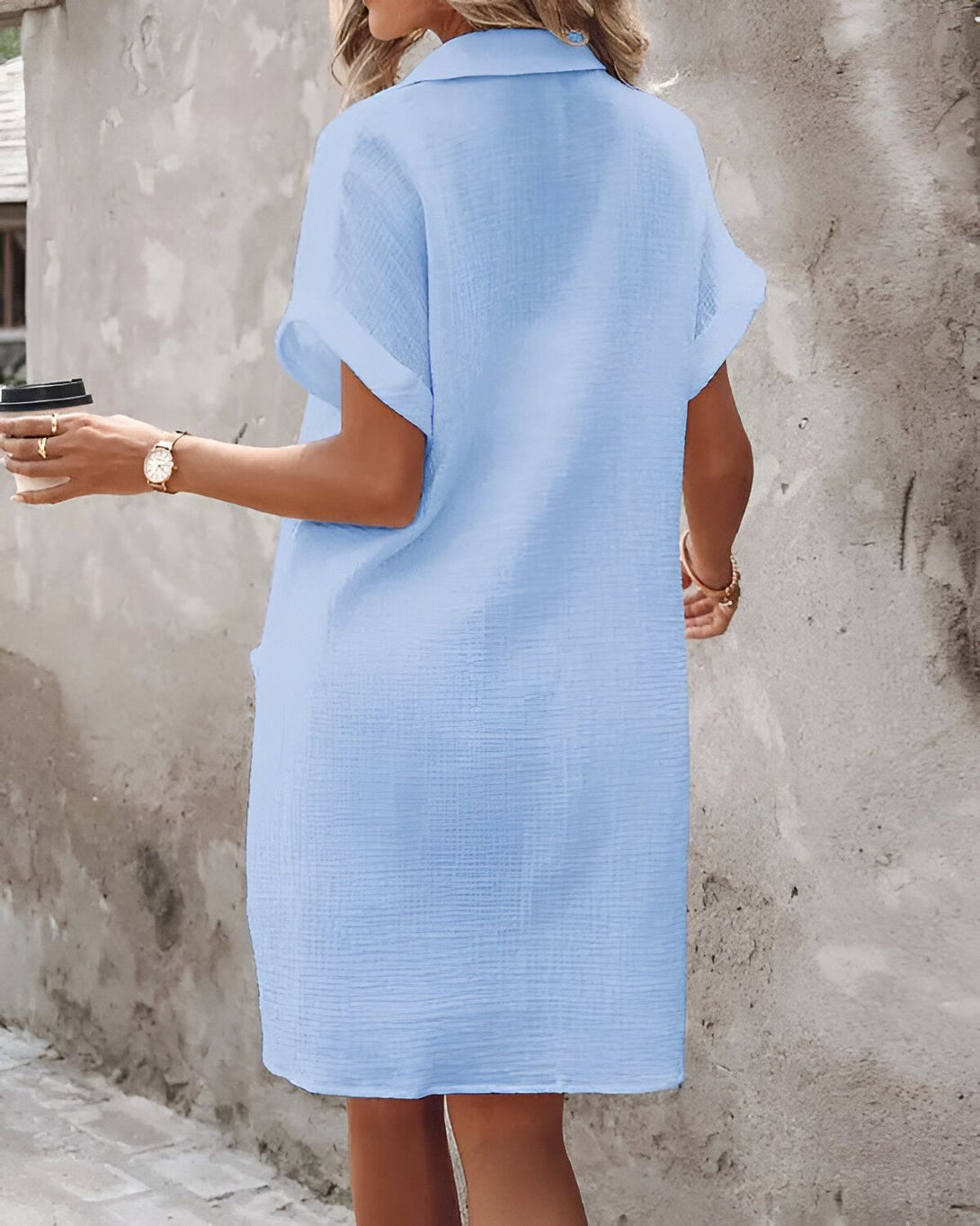 Une femme porte une robe chemise bleu clair mi-longue à manches courtes. Elle est vue de dos.