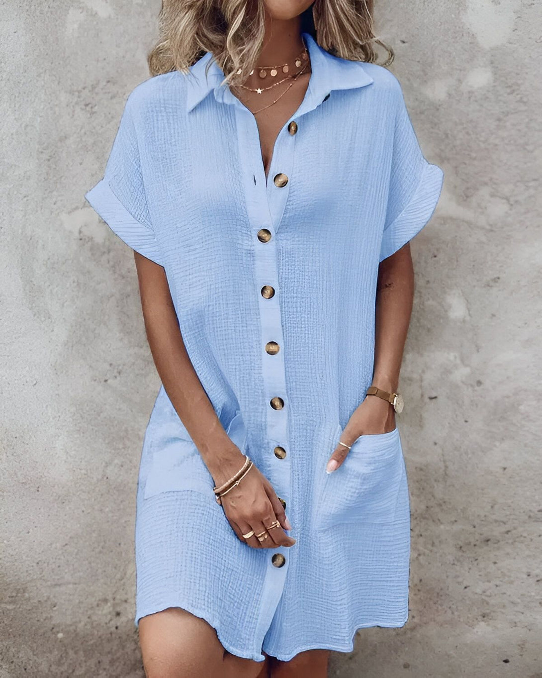 Une jeune femme porte une chemise ample bleu clair à manches courtes. Elle est ouverte sur le devant et possède des boutons. La femme a les cheveux blonds et porte un collier. Elle se tient debout, les mains dans les poches.