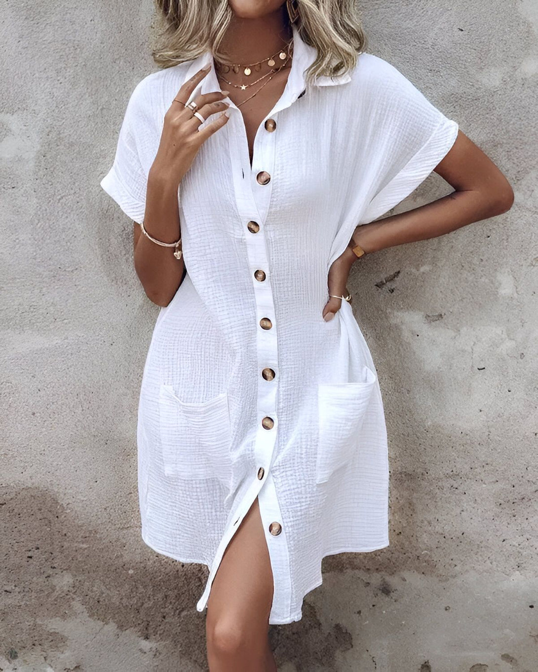 Une jeune femme porte une robe chemise blanche courte à manches courtes. La robe est ouverte sur le devant et on peut voir un bouton doré. La femme porte également un collier doré et des bracelets. Elle a les cheveux blonds et le maquillage léger.