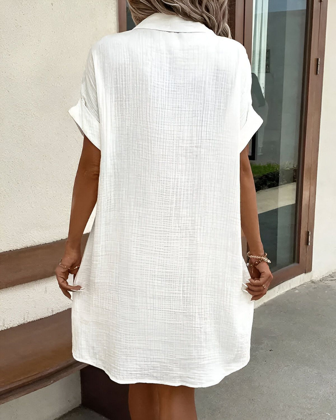 Une femme porte une robe chemise blanche courte à manches courtes. Elle est vue de dos.