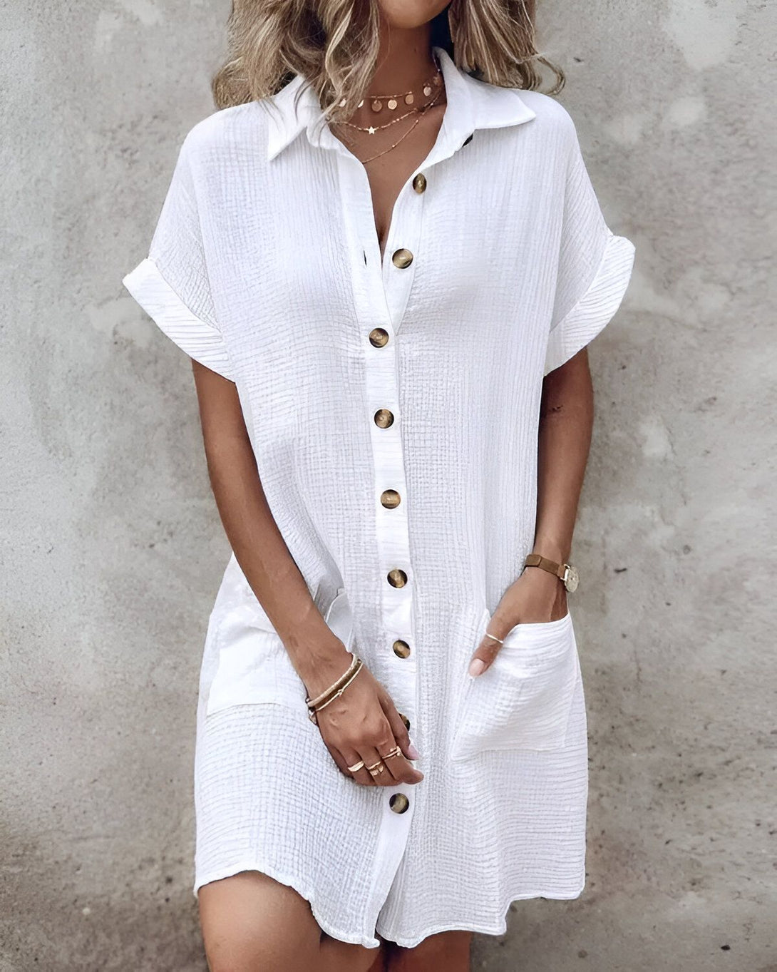 Une jeune femme porte une robe chemise blanche en lin. La robe est ouverte sur le devant et possède des boutons. La femme a les cheveux blonds et porte un collier. Elle est debout devant un mur gris.