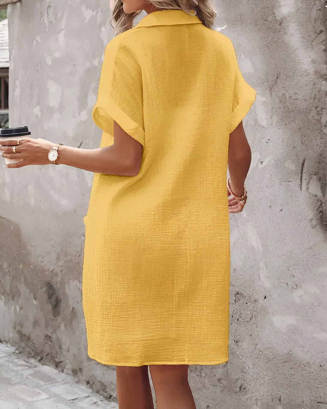 Une femme porte une robe jaune courte à manches courtes et un col montant. Elle tient une tasse de café à la main.