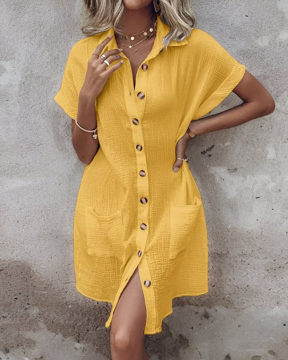 Une jeune femme porte une robe chemise jaune boutonnée. La robe est courte et possède des manches courtes. La femme est blonde et porte un collier. Elle est souriante et regarde l'appareil photo.