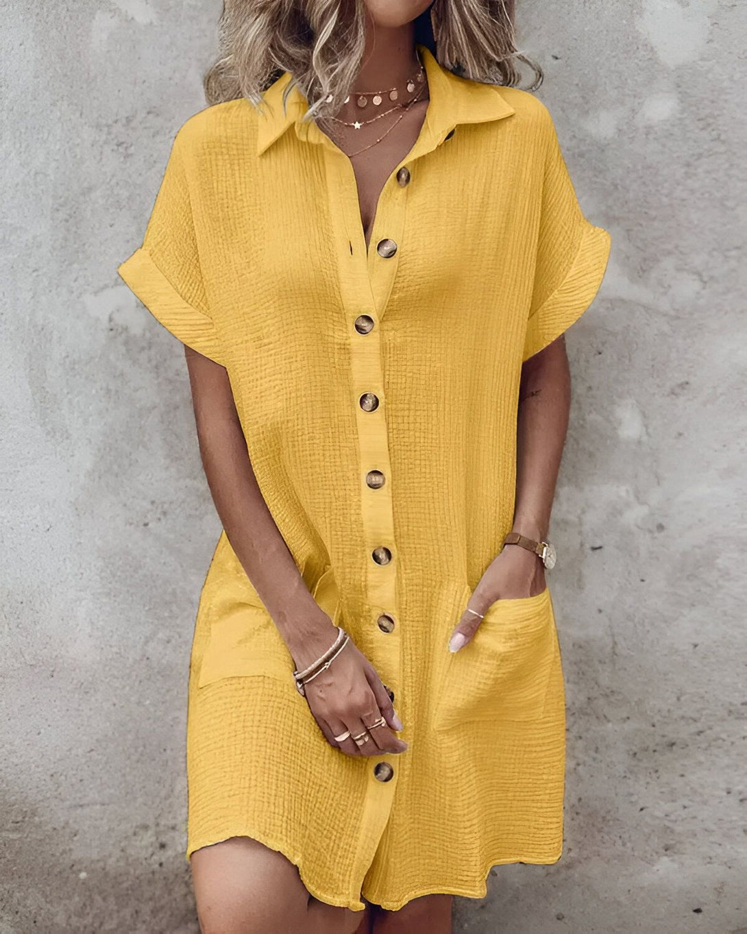 Une femme porte une robe chemise jaune boutonnée. La robe est courte et a des manches courtes. La femme a les cheveux longs et blonds. Elle porte un collier et des bracelets. Elle est debout devant un mur gris.