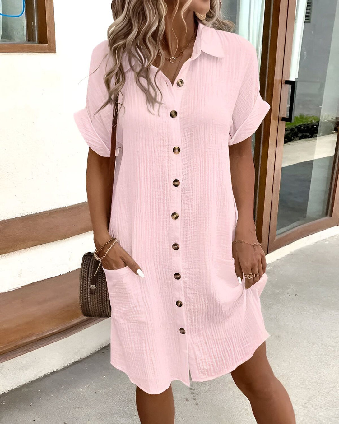 Une femme porte une robe chemise rose boutonnée. Elle a les cheveux longs et blonds. Elle est debout devant une porte vitrée.