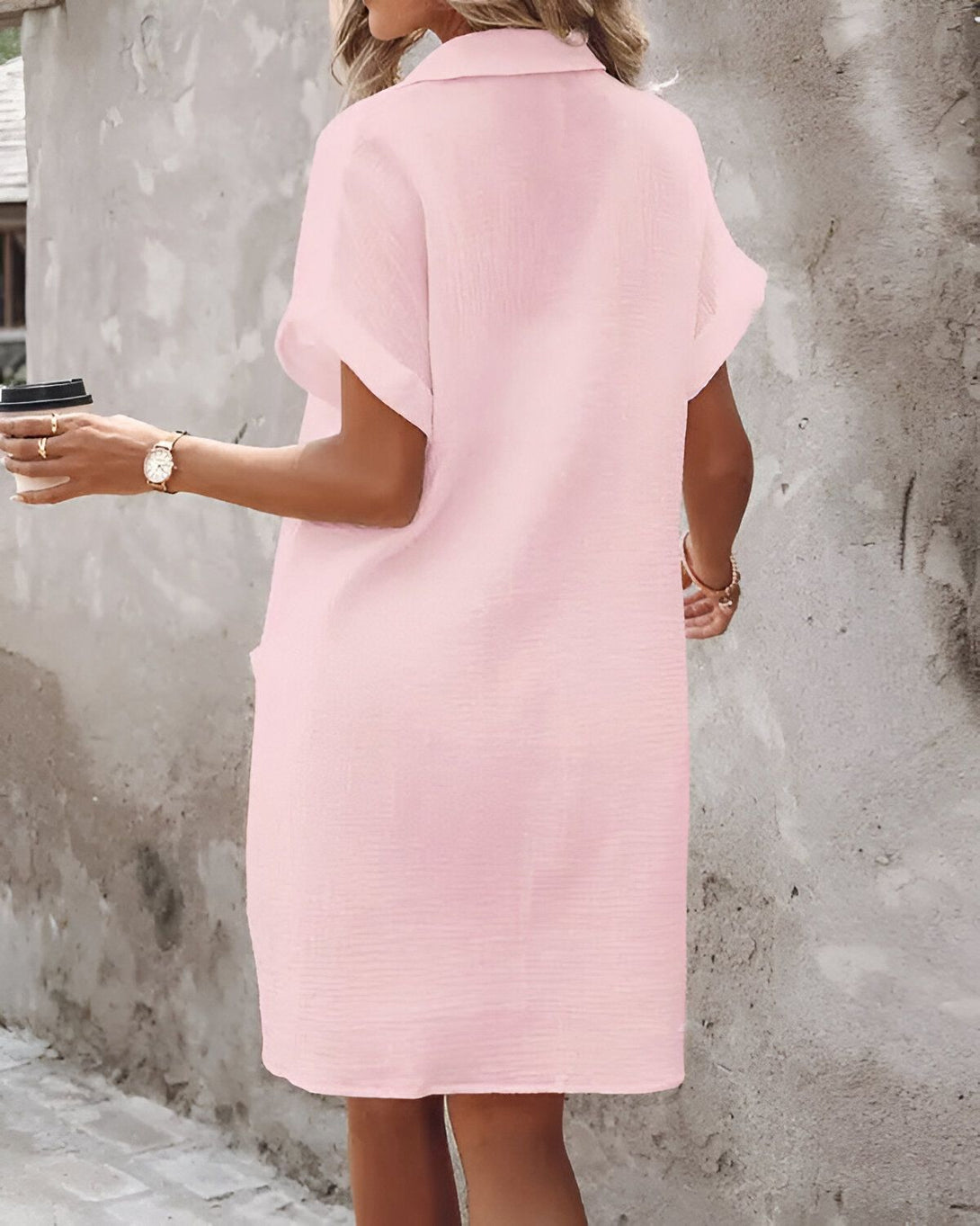 Une femme porte une robe chemise rose. Elle a les cheveux longs et bruns et porte un sac à main marron. Elle est en train de marcher dans la rue.
