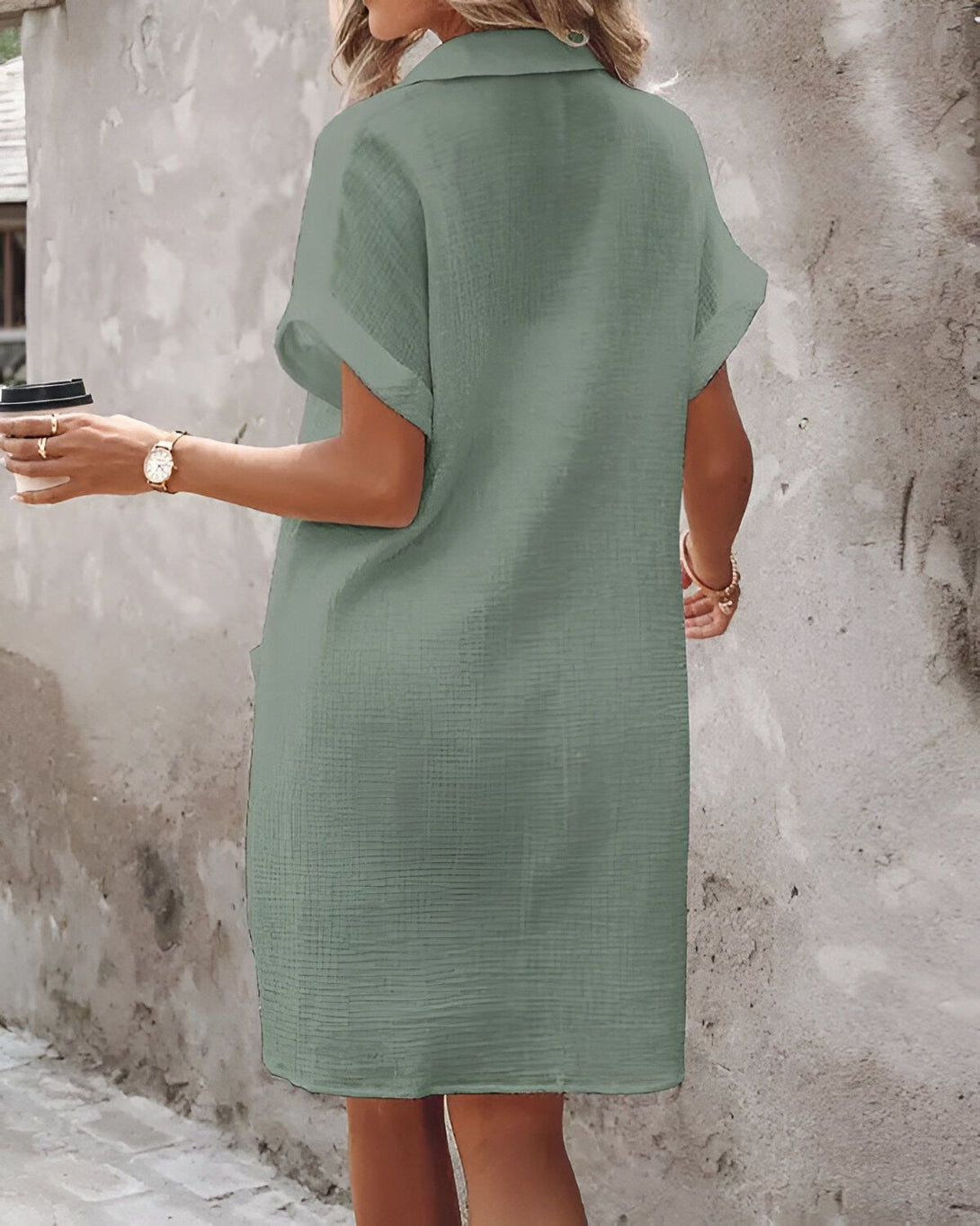 Une femme porte une robe chemise verte courte à manches retroussées. Elle a une montre au poignet gauche et tient une tasse à café dans sa main droite.