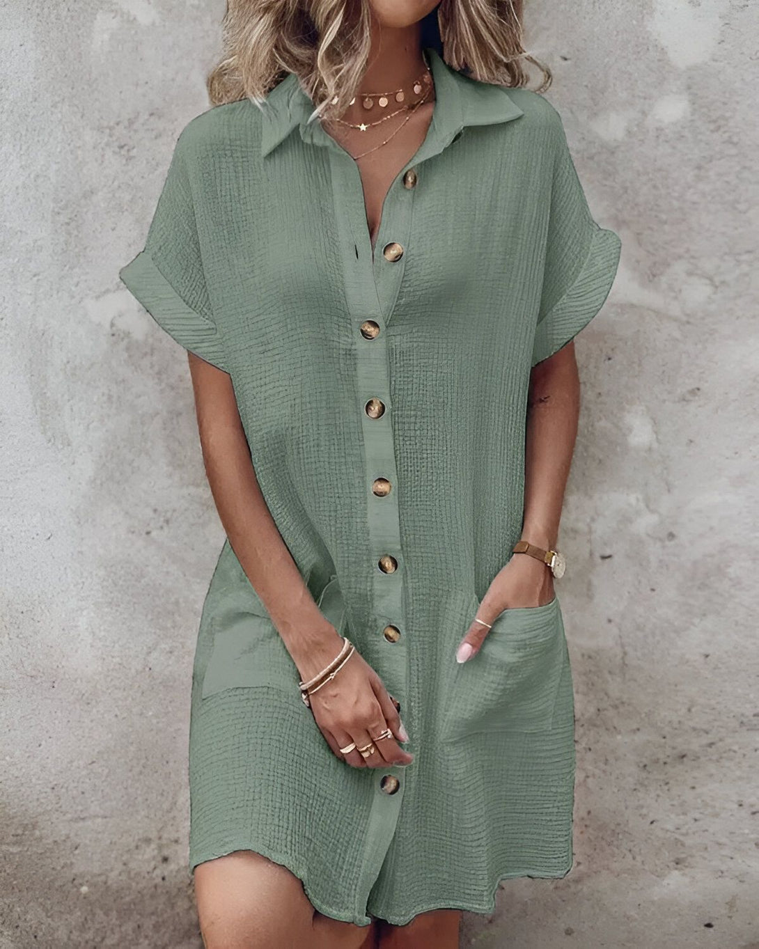 Une femme porte une robe chemise verte courte à manches courtes. La robe est ouverte sur le devant et on peut voir un t-shirt blanc en dessous. La femme a une main dans la poche de la robe et l'autre tient le bas de la robe. Elle porte un collier et des bracelets.