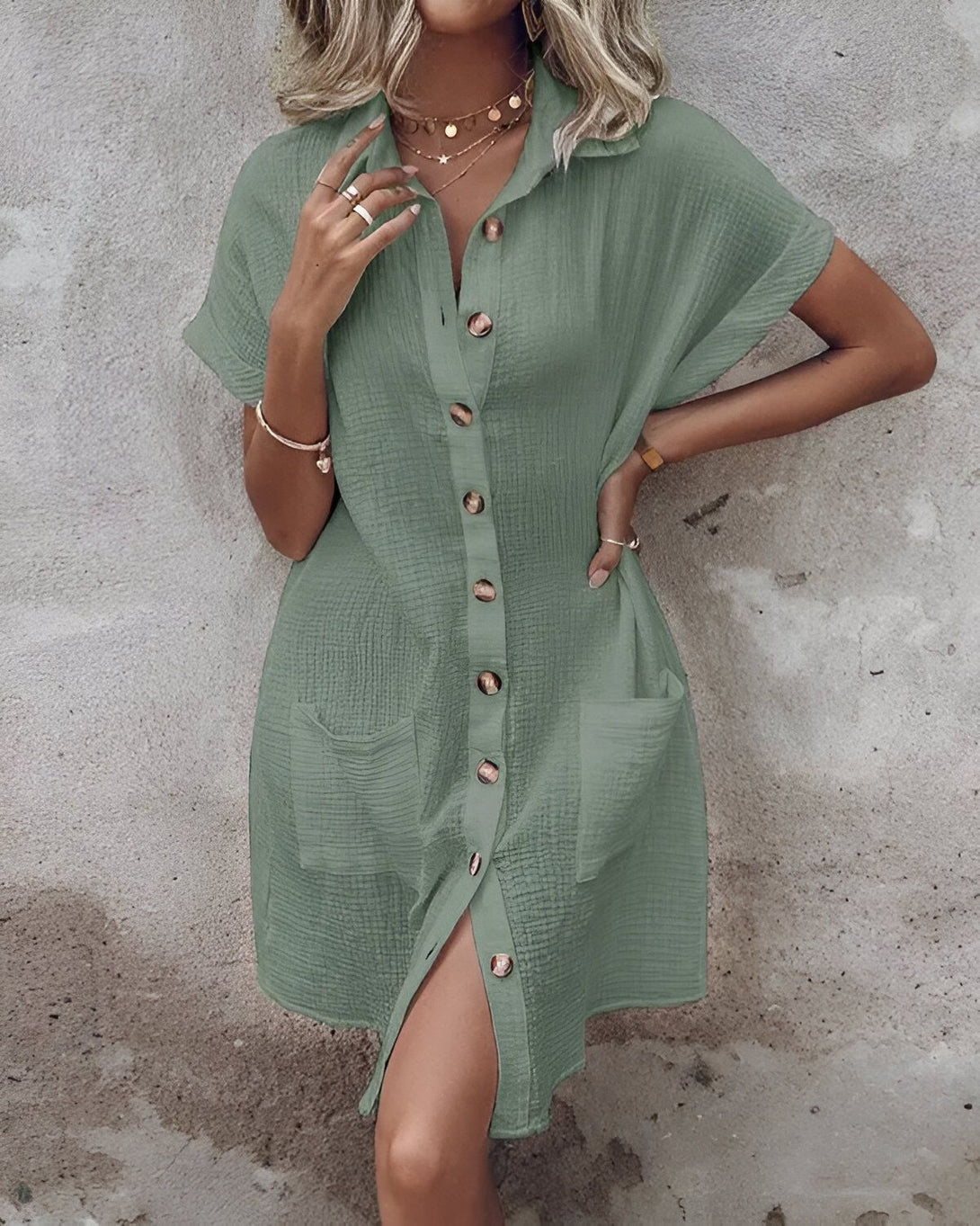 Une jeune femme porte une robe chemise verte courte à manches courtes. La robe est ouverte sur le devant et l'on peut voir un t-shirt blanc en dessous. La femme est blonde et porte un collier avec un pendentif en forme d'étoile. Elle a également un bracelet au poignet gauche.