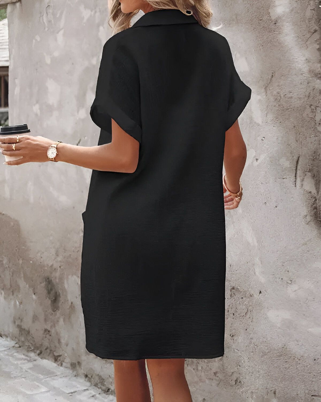 Une femme porte une robe noire décontractée. Elle est vue de dos. Ses cheveux sont longs et blonds. Elle porte une montre au poignet gauche et tient une tasse de café à la main droite.