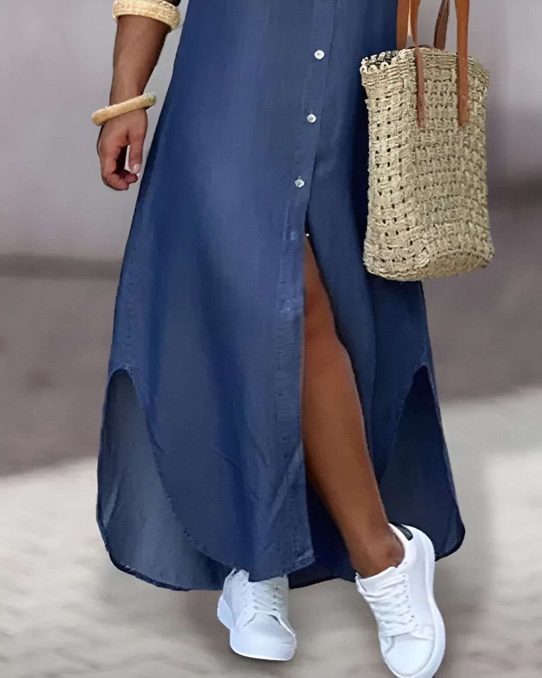 Une femme porte une longue robe chemise en jean bleu avec des boutons sur le devant. Elle est fendue sur les côtés et laisse entrevoir ses jambes. Elle porte également des baskets blanches et un sac à main en paille marron.