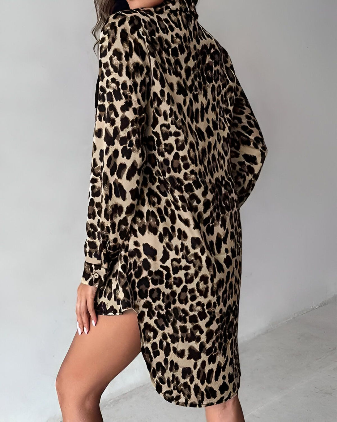 Une femme porte une chemise léopard oversize avec des boutons marron. La chemise est ouverte sur le bas et on peut voir ses jambes. Elle a les cheveux longs et bruns et porte un bracelet à la main droite.