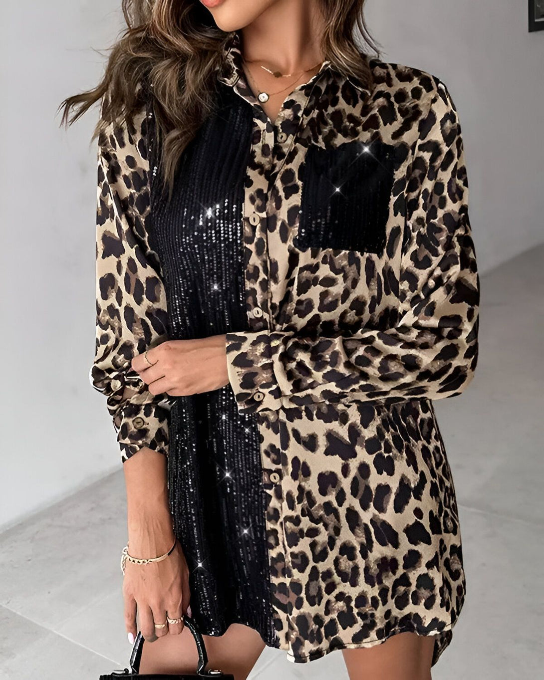 Une femme porte une chemise léopard avec des sequins noirs. La chemise est ouverte et on peut voir un t-shirt noir en dessous. La femme a les cheveux longs et bruns et elle porte un collier. Elle est debout dans un magasin et il y a des vêtements sur les étagères derrière elle.