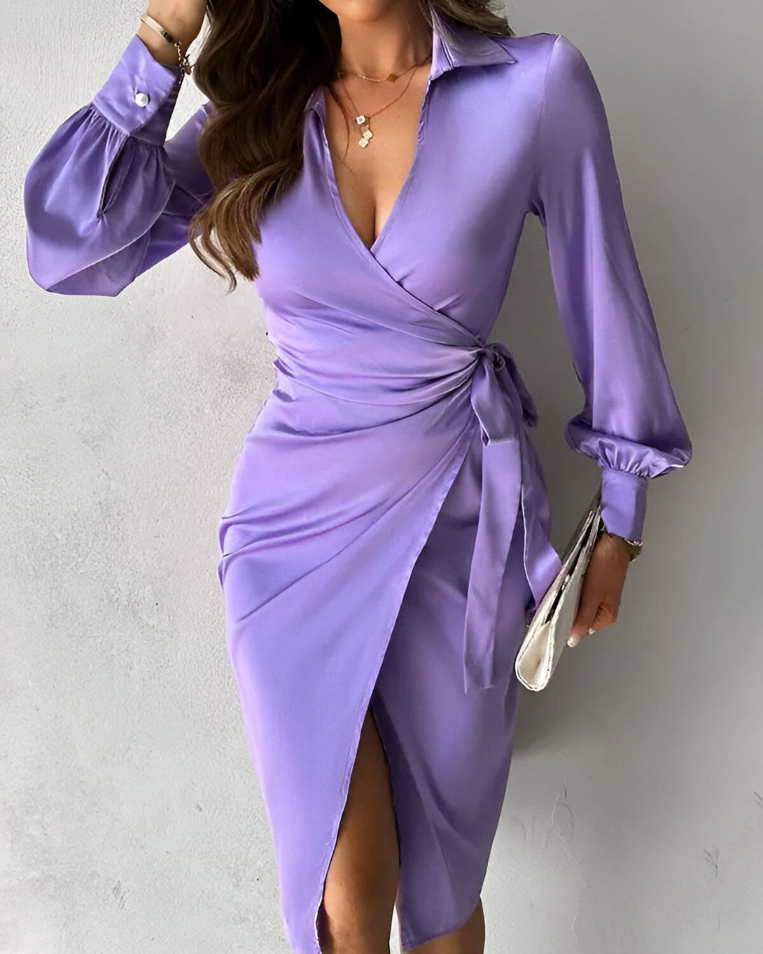 Une femme porte une robe portefeuille en satin lavande. La robe a un décolleté en V profond et des manches longues bouffantes. Elle est ceinturée à la taille et possède une fente sur le côté. La femme est debout, les cheveux détachés et porte un collier.