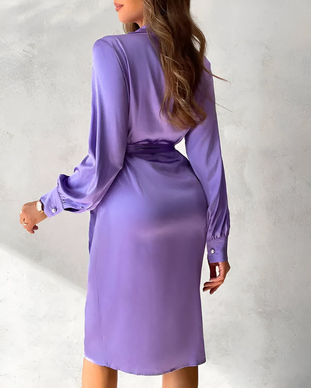 Une femme porte une robe chemise en satin mauve. La robe est ceinturée à la taille et possède un col montant. Les manches sont longues et bouffantes. La femme est vue de dos.