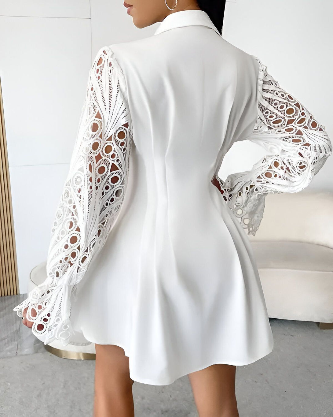 Une femme porte une robe blanche en dentelle. La robe est cintrée à la taille et évasée au niveau des hanches. Elle a des manches longues et un col montant. La femme est vue de dos.