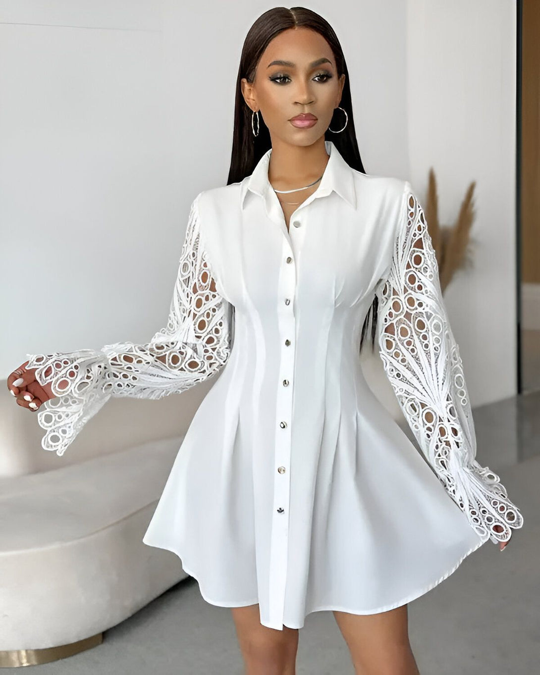 Une jeune femme porte une robe chemise blanche. La robe est cintrée à la taille et évasée au niveau des hanches. Elle a des manches longues et un col montant. La femme est debout, les bras légèrement écartés du corps. Elle a les cheveux longs et noirs et porte un maquillage léger.