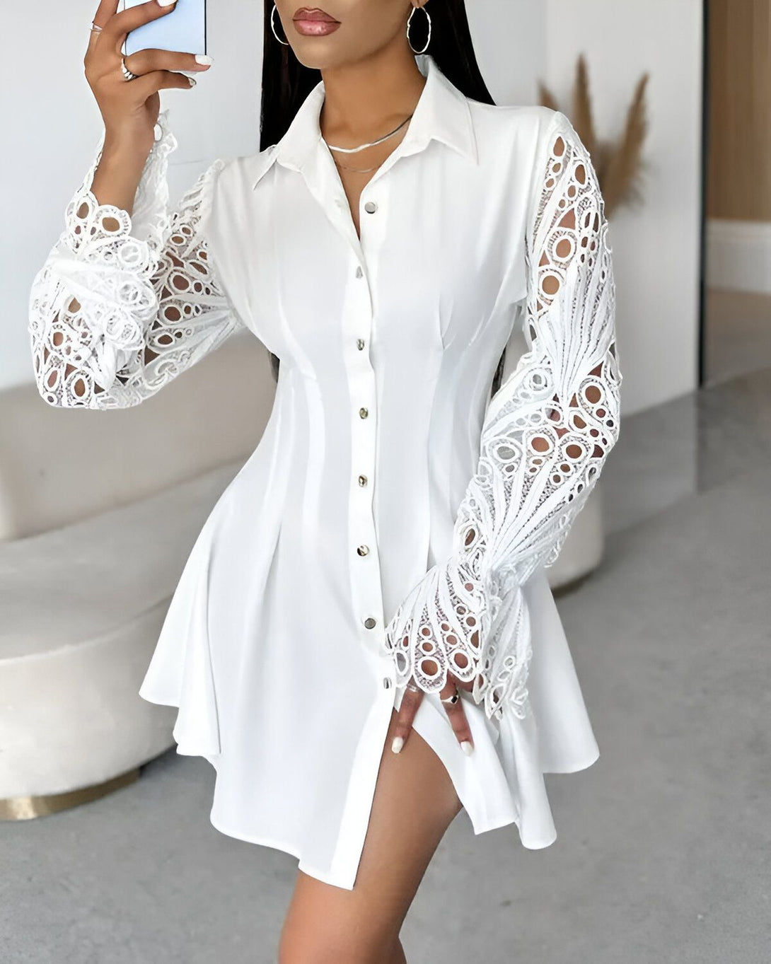 Une femme porte une robe chemise blanche. La robe est cintrée à la taille et évasée au niveau des hanches. Elle a des manches longues en dentelle. La femme est debout, elle a une main sur la hanche et l'autre tient un téléphone portable.