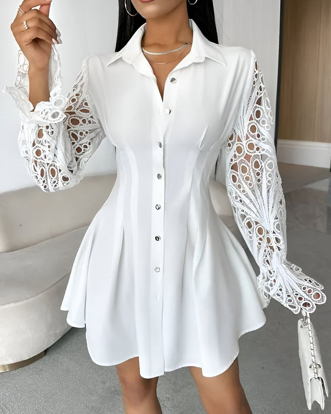 Une jeune femme porte une robe chemise blanche. La robe est cintrée à la taille et évasée au niveau des hanches. Elle a des manches longues en dentelle. La femme est debout, elle a la main sur la hanche et regarde l'objectif. Elle porte un collier et des boucles d'oreilles.