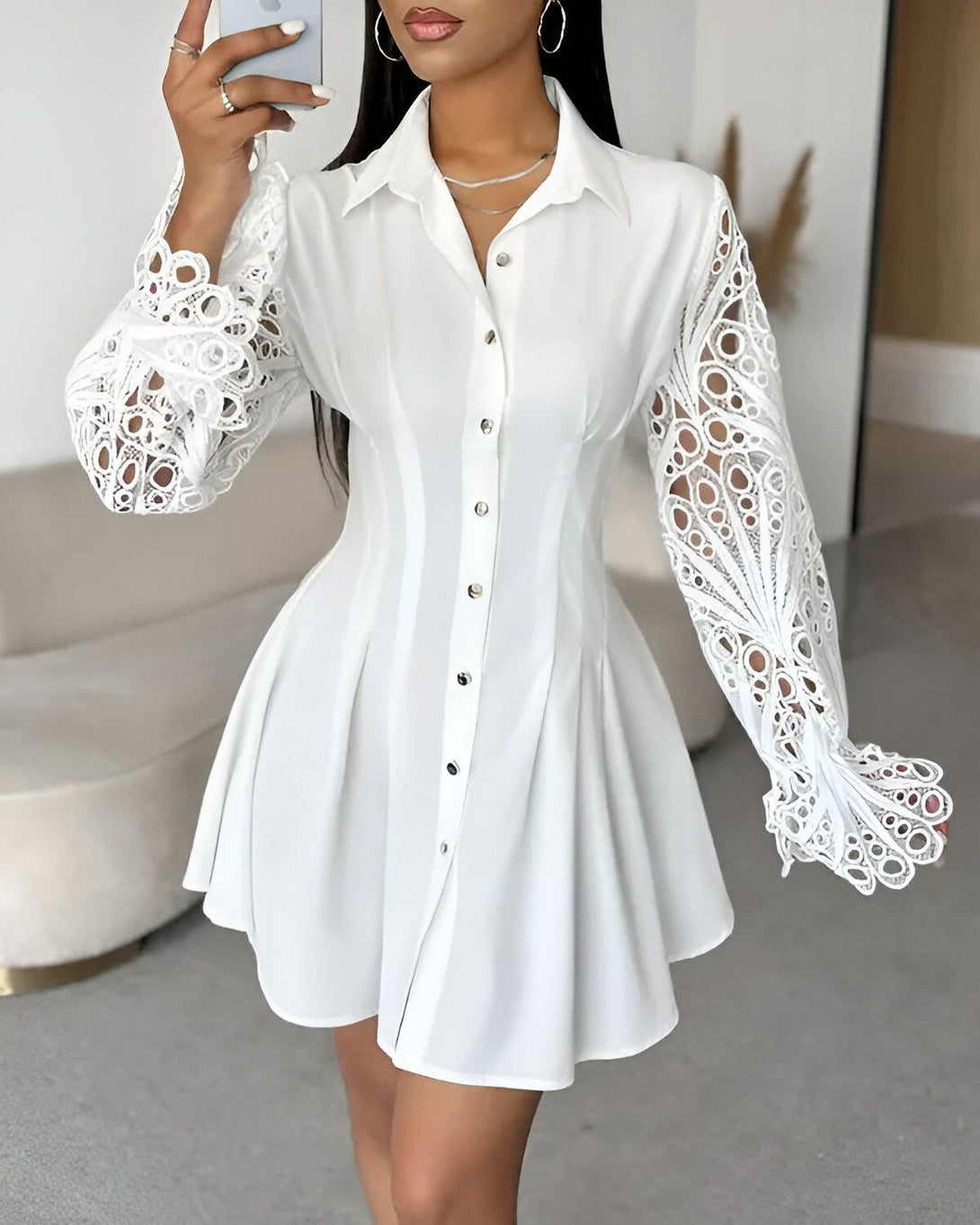 Une femme porte une robe chemise blanche. La robe est cintrée à la taille et évasée au niveau des hanches. Elle a des manches longues et un col montant. La femme est debout, elle a les cheveux longs et noirs et elle porte du maquillage.