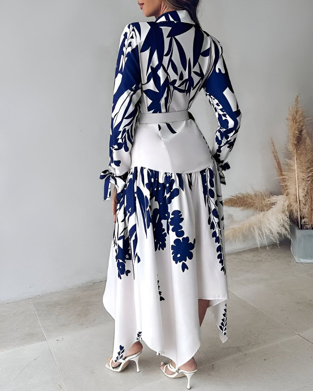 Une femme porte une robe longue blanche et bleue à motifs floraux. La robe est cintrée à la taille et possède un col montant. Les manches sont longues et bouffantes. La jupe est évasée et fendue sur le côté. La femme porte des escarpins blancs et un collier bleu. Elle est debout dans un intérieur, adossée à un mur blanc.