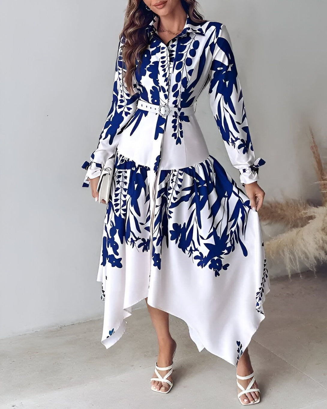 Une femme porte une robe chemise longue bleu et blanche à motifs floraux. La robe est ceinturée à la taille et possède un col montant. Les manches sont longues et bouffantes. La femme porte des escarpins blancs et un sac à main blanc.