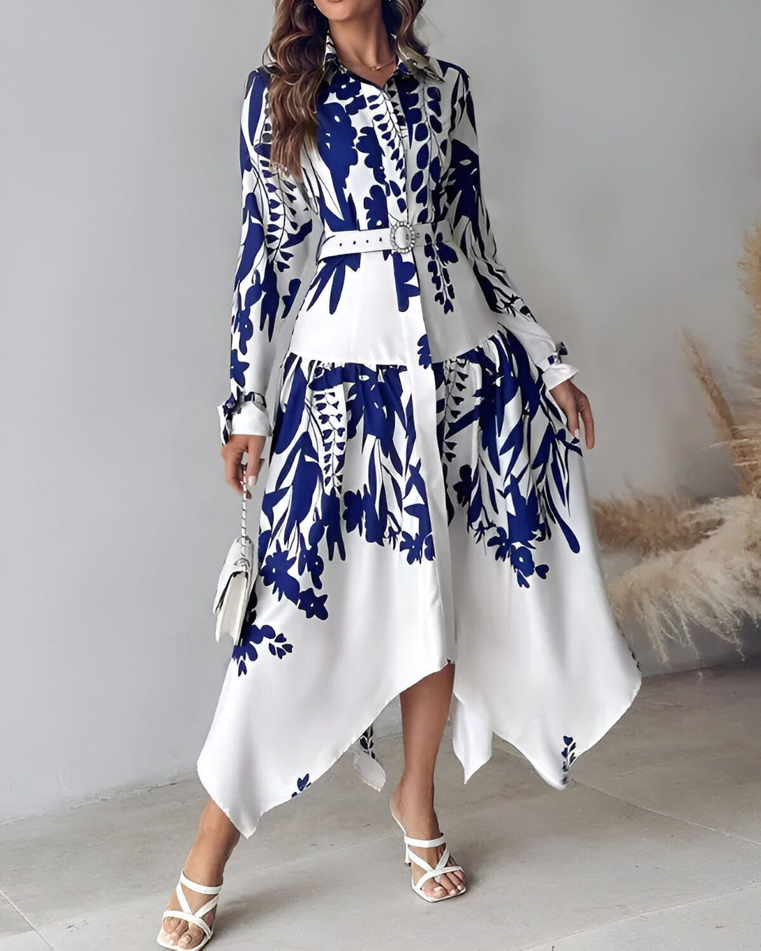 Une femme porte une robe chemise longue à imprimé floral bleu et blanc. La robe est cintrée à la taille et possède un ourlet asymétrique. La femme porte des sandales à talons blanches et un sac à main blanc.