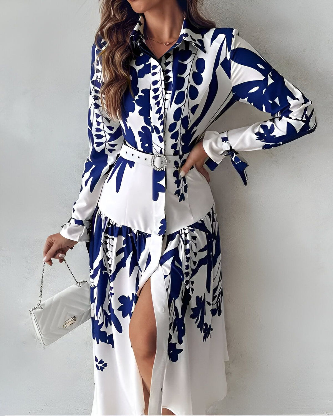 Une femme porte une robe chemise longue à imprimé floral bleu et blanc. La robe est ouverte sur le devant et nouée à la taille par une ceinture. La femme porte également un sac à main blanc.