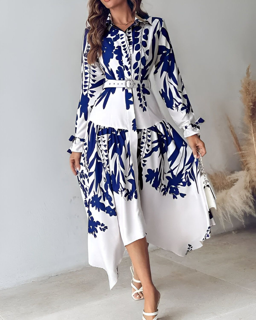Une femme porte une robe chemise longue blanche à motifs bleus. La robe est ceinturée à la taille et possède un col montant. Les manches sont longues et bouffantes. La femme porte des sandales à talons blanches et un sac à main blanc.