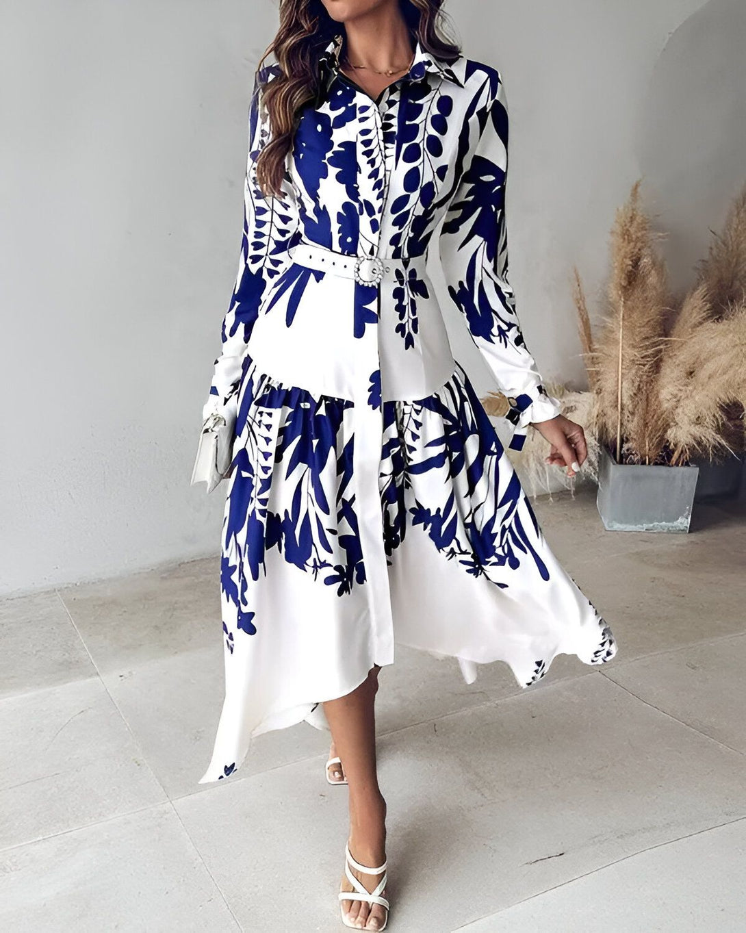 Une femme porte une robe chemise longue blanche à motifs bleus. La robe est ceinturée à la taille et possède un col montant. Les manches sont longues et bouffantes. La femme porte des sandales à talons blanches.