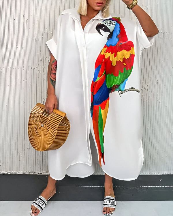 Une femme porte une longue chemise blanche avec un perroquet coloré imprimé dessus. Elle porte également un sac à main marron et des sandales blanches.