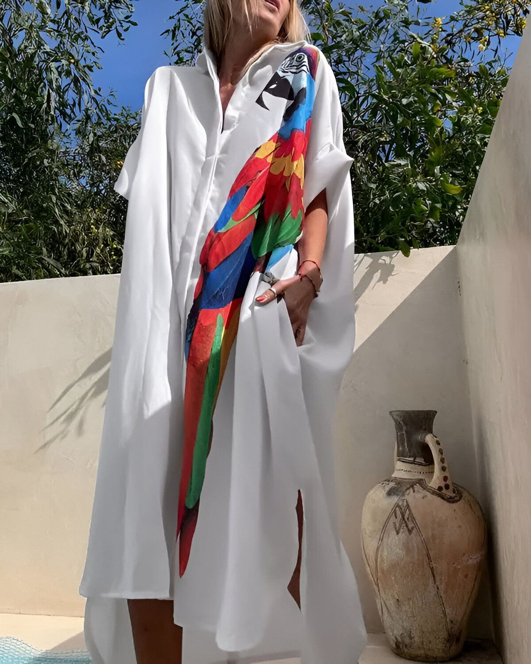 Une femme porte une longue chemise blanche avec un perroquet coloré imprimé dessus. Elle se tient debout sur une terrasse, avec des arbres et un ciel bleu en arrière-plan.