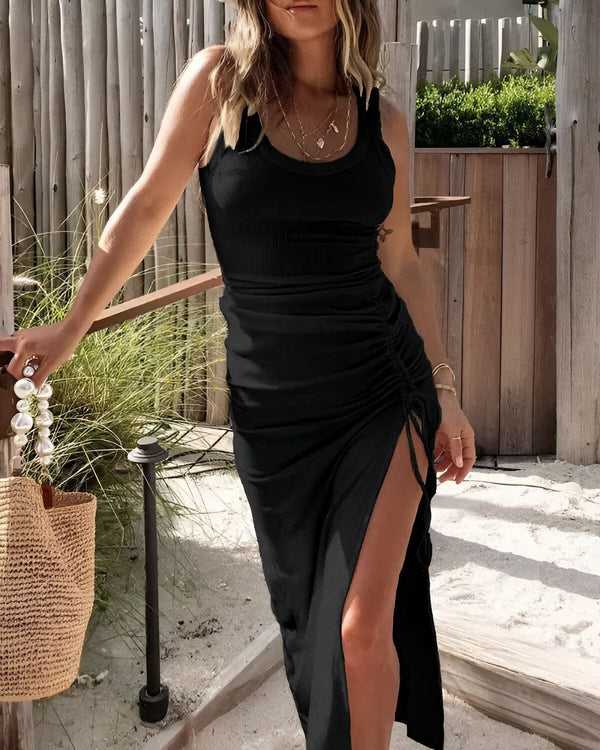 Une femme porte une robe noire moulante avec une fente sur le côté. Elle est debout sur une terrasse et tient un sac à main en osier.