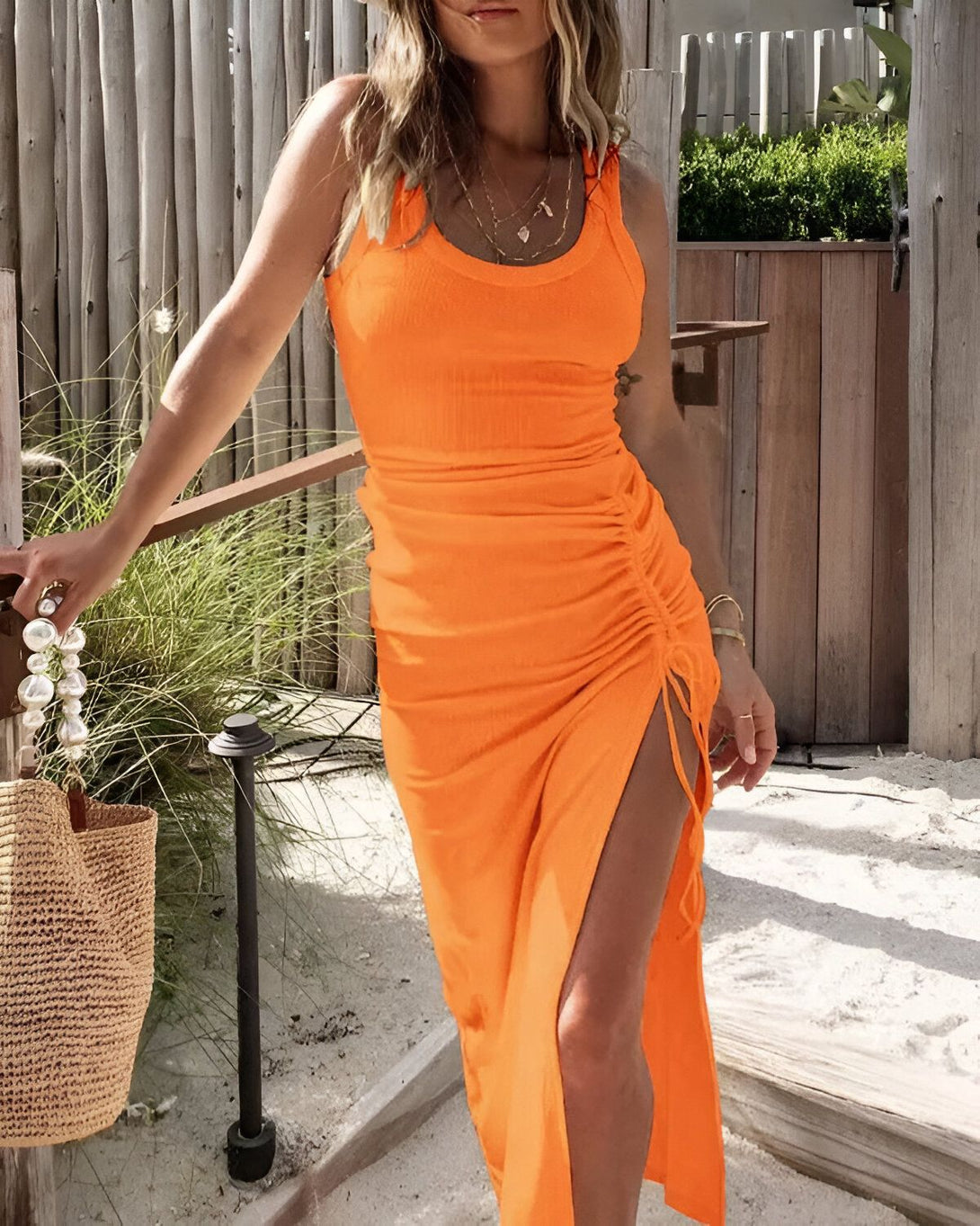Une femme porte une robe orange moulante avec une fente sur le côté. Elle a les cheveux longs et blonds et porte un collier de perles. Elle est debout sur une terrasse en bois et tient un sac à main en osier à la main.