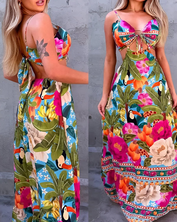 Une femme porte une robe longue à fines bretelles. La robe est ouverte dans le dos et nouée au niveau de la taille. Elle est imprimée de fleurs tropicales colorées et de feuilles vertes. La femme est blonde et porte un maquillage léger. Elle a les cheveux longs et lâchés. Elle est pieds nus.