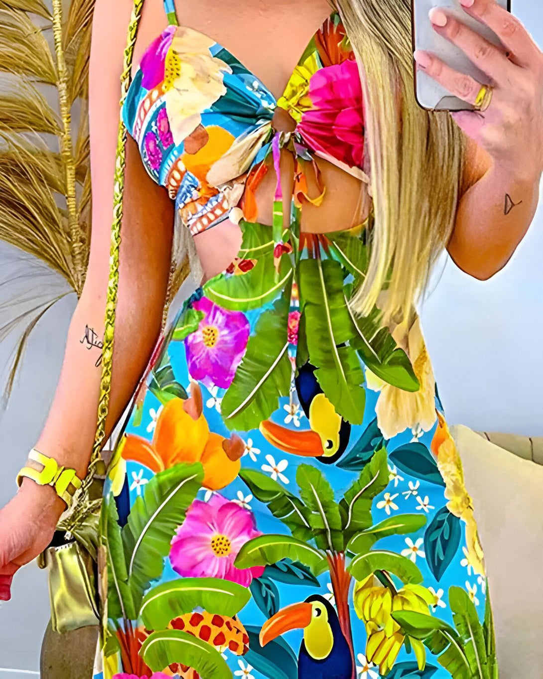 Une femme porte une robe d'été colorée avec des fleurs et des feuilles tropicales. Elle est debout dans une pose avantageuse et tient un téléphone portable à la main.
