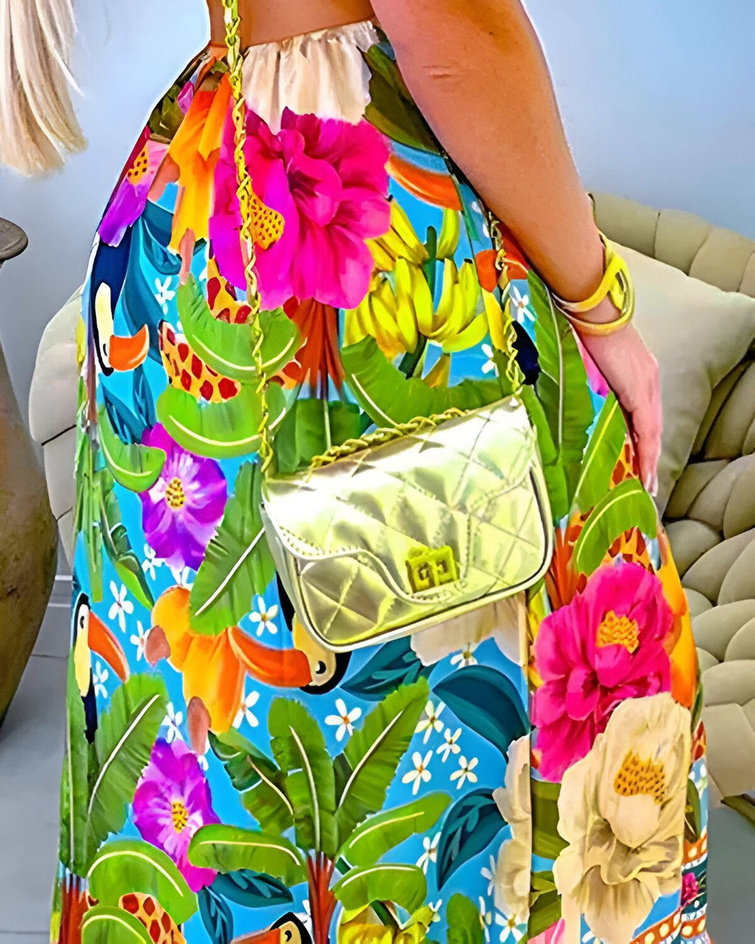 Une femme porte une robe longue colorée à motifs tropicaux et un sac à main doré.