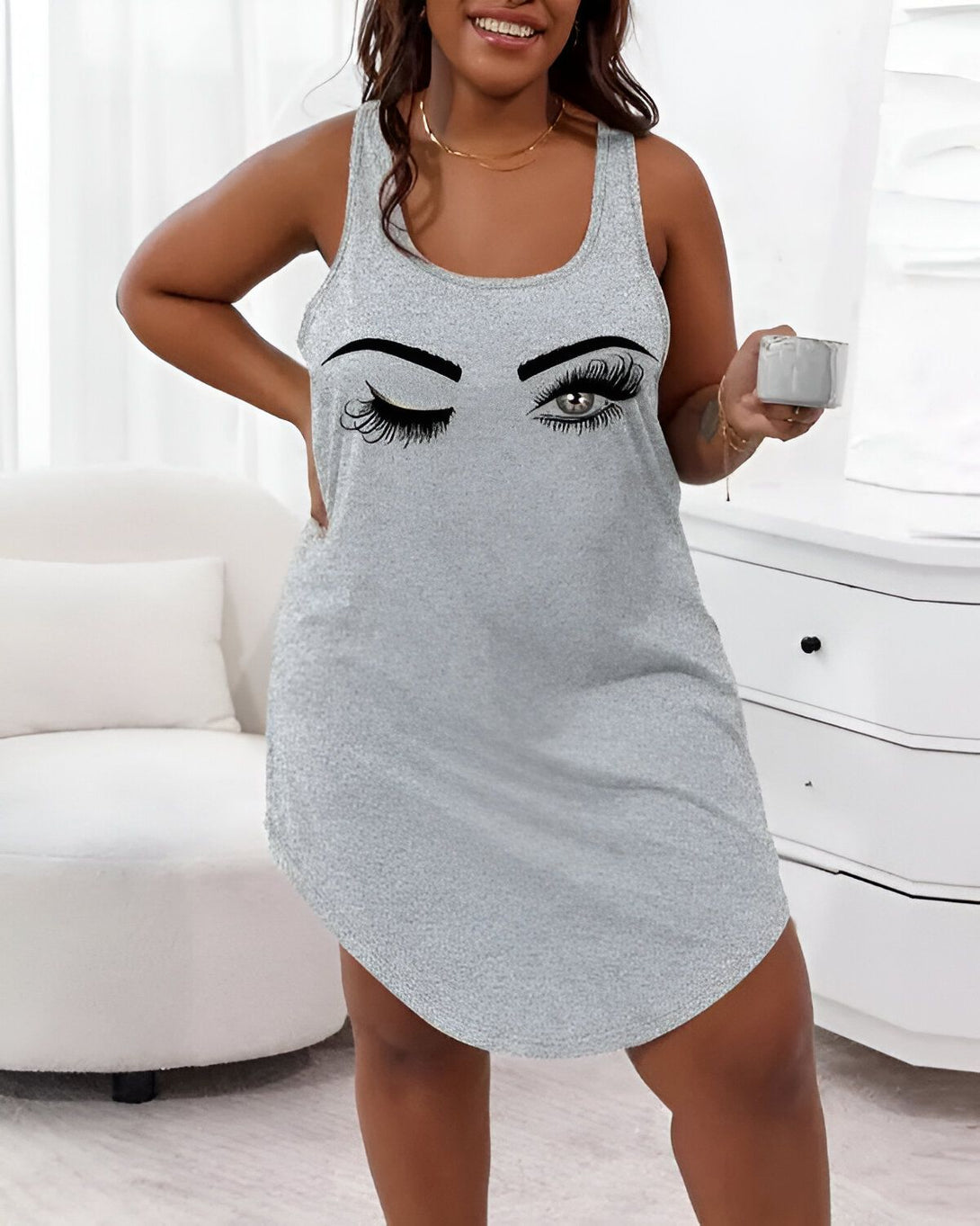 Une femme porte une nuisette grise avec un dessin d'un clin d'œil sur la poitrine. Elle est debout dans une chambre et tient une tasse à la main.