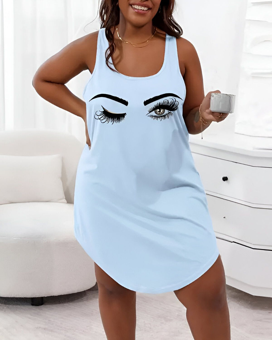 Une femme porte une nuisette bleue avec un dessin d'un clin d'œil sur la poitrine. Elle est debout dans une chambre et tient une tasse à la main.