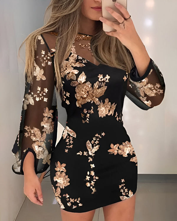 Une femme porte une robe noire avec des fleurs dorées. La robe est moulante et a des manches longues. La femme est debout dans un miroir et se prend en photo.