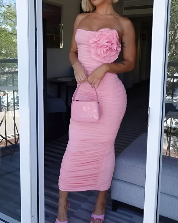 Une femme porte une robe rose moulante sans bretelles avec une fleur rose sur le côté. Elle porte également des chaussures à talons roses et un sac à main rose.