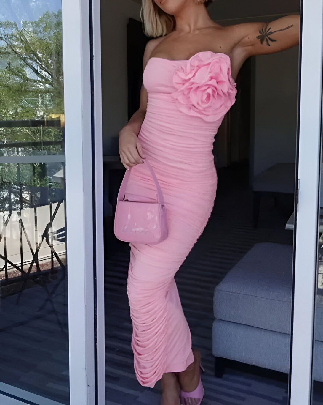 Une femme porte une robe rose moulante sans bretelles avec une fleur rose sur le côté. Elle porte également des sandales à talons roses et un sac à main rose.