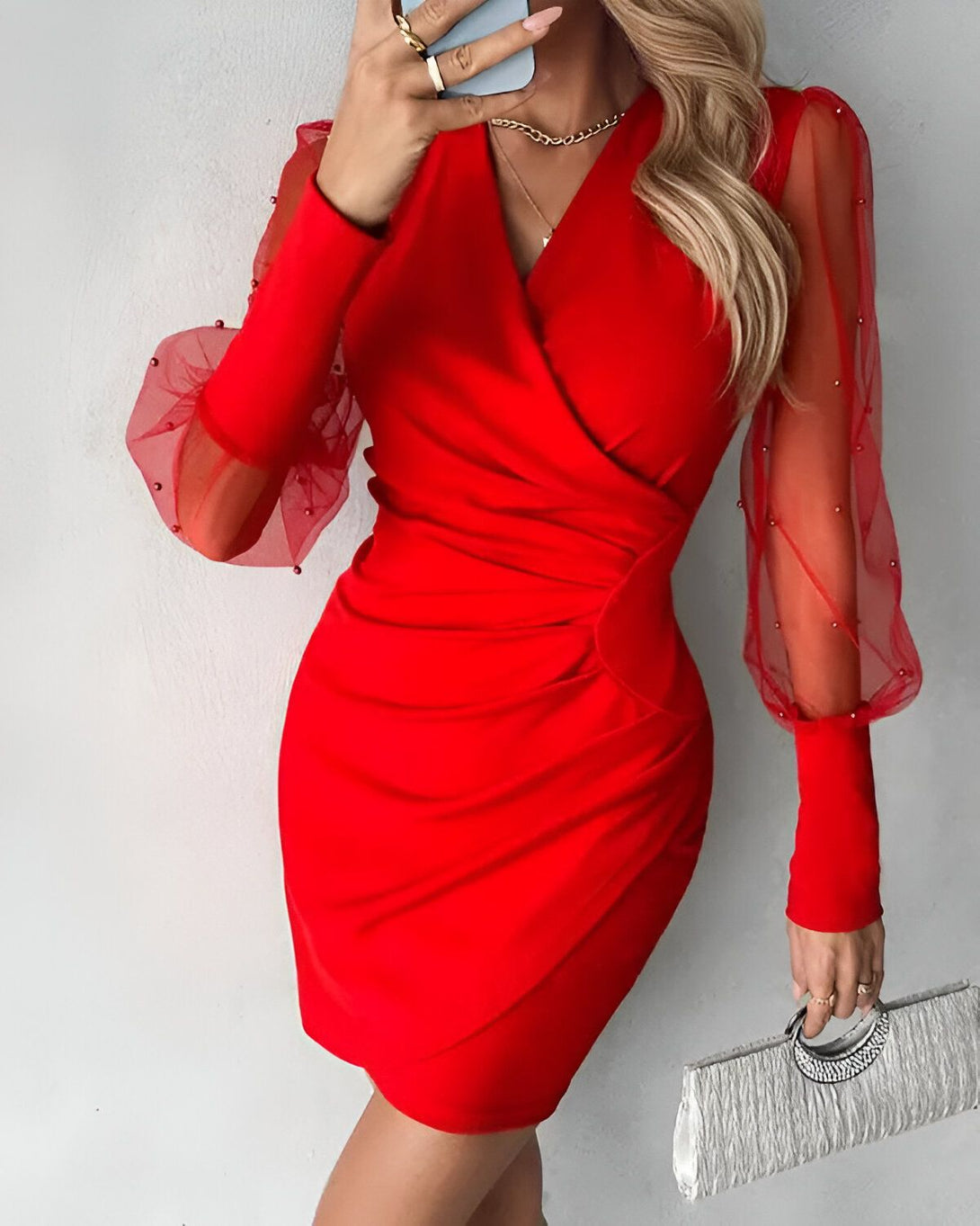 Une femme porte une robe rouge moulante avec des manches bouffantes en tulle perlé. Elle est également porte une pochette argentée et un collier doré.