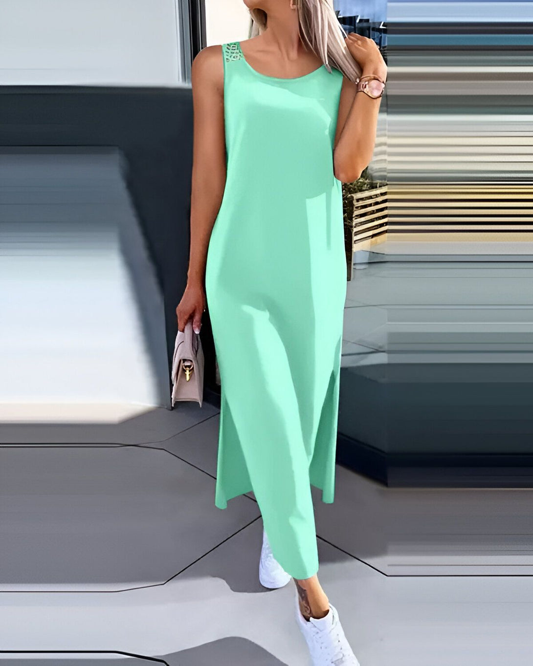 Une femme porte une robe longue vert menthe. La robe est sans manches et a un col rond. Elle est fendue sur les côtés. La femme porte des baskets blanches et un sac à main beige.