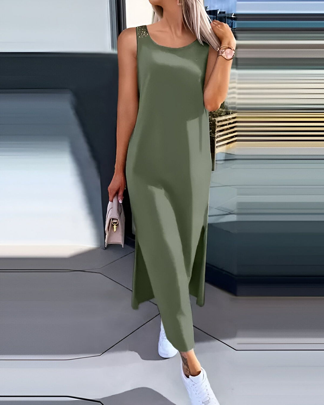 Une femme porte une robe longue vert kaki sans manches. La robe est fendue sur les côtés. Elle porte des baskets blanches et un sac à main beige.
