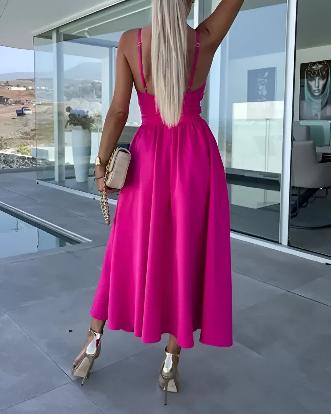 Une femme blonde porte une robe longue rose dos nu. Elle est de dos et porte des sandales à talons hauts couleur or. Elle porte un sac à main beige à la main droite.