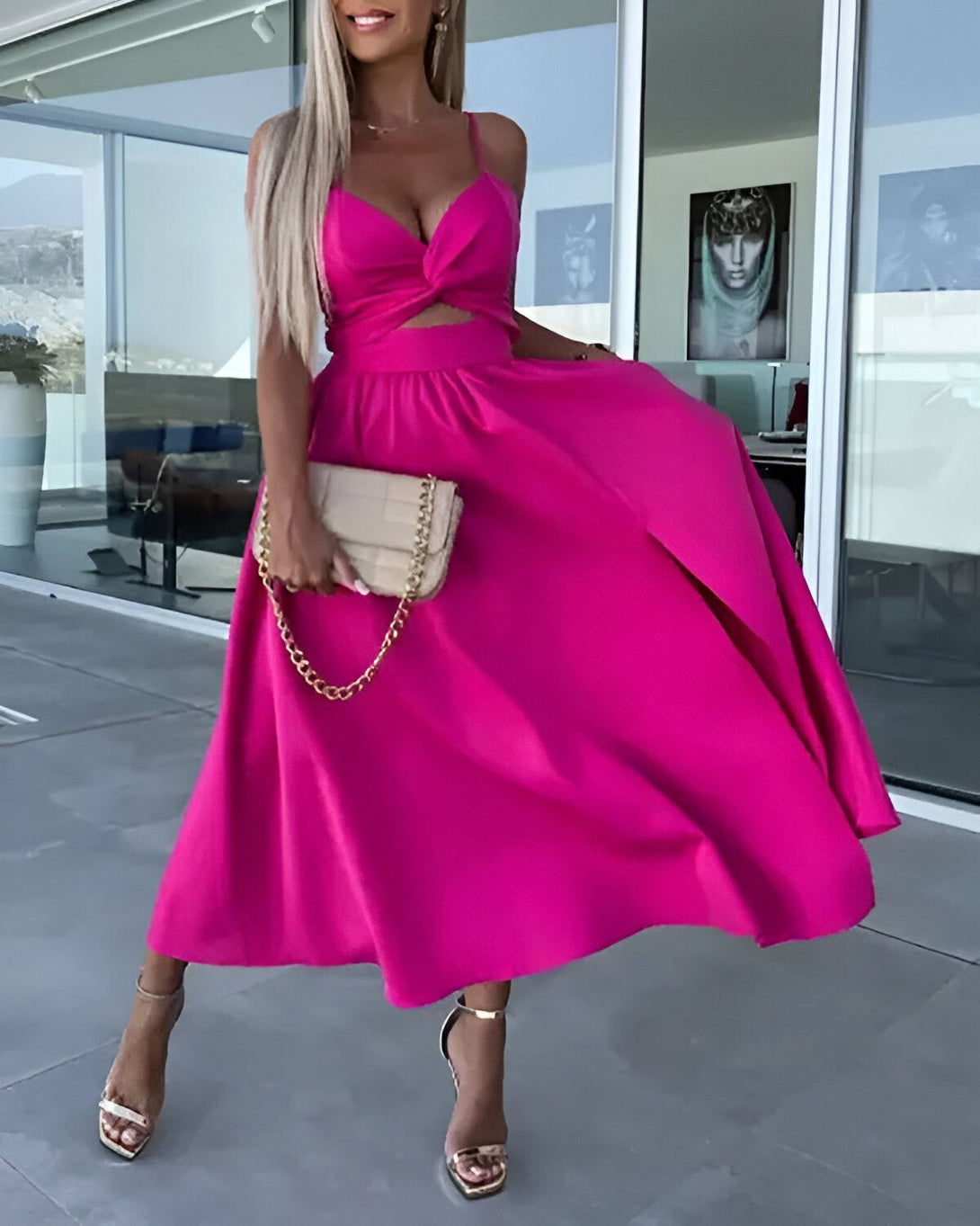 Une femme porte une robe longue rose fendue sur le côté. Elle est décolletée et a un nœud au niveau de la poitrine. Elle porte des sandales à talons hauts et un sac à main beige.