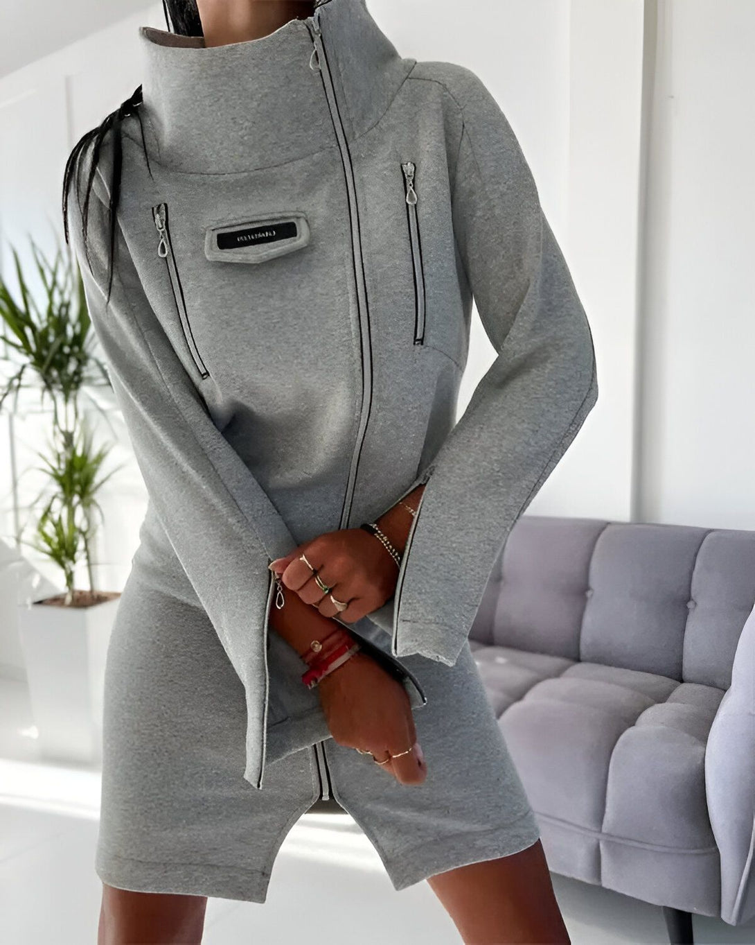 Une femme porte une robe sweat-shirt grise à fermeture éclair. La robe est courte et possède un col montant. Les manches sont longues et évasées. La femme est debout dans un salon, adossée à un canapé gris.