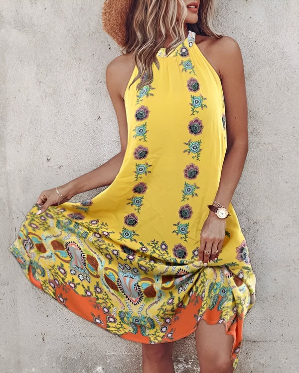 Une femme porte une robe jaune à motifs floraux. La robe est courte et évasée, avec un col montant et des bretelles fines. La femme est debout, les cheveux longs et blonds lâchés. Elle porte un chapeau de paille marron et des sandales jaunes.