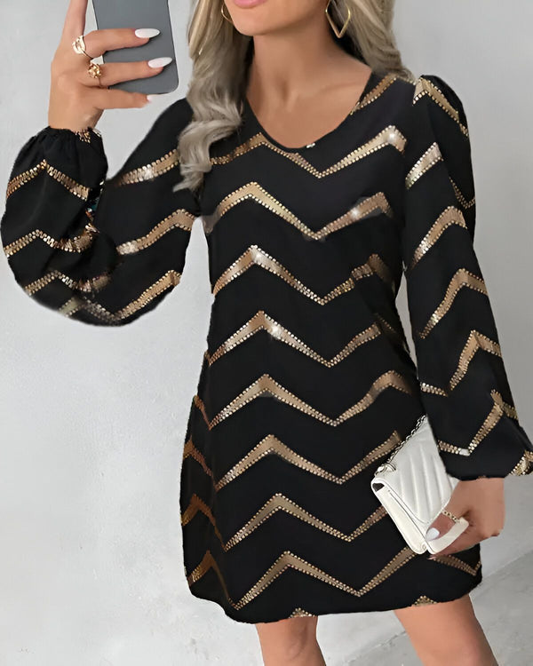 Une femme porte une robe noire à sequins dorés. La robe est à manches longues et a une encolure ronde. La femme tient un téléphone portable dans sa main droite et un sac à main blanc dans sa main gauche.